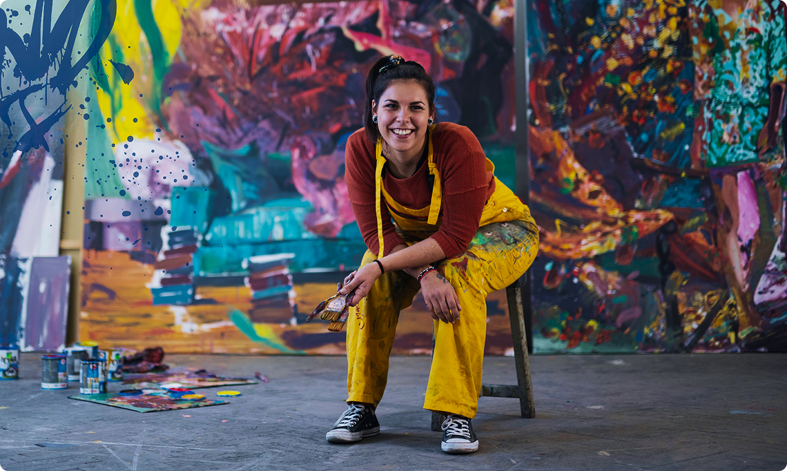 jovem suja de tinta vestida de macacao branco sujo de tinta, ela est&aacute; sentada em uma cadeira olhando para a frente, atras dela tem a obra que ela pintou e ela est&aacute; alegre